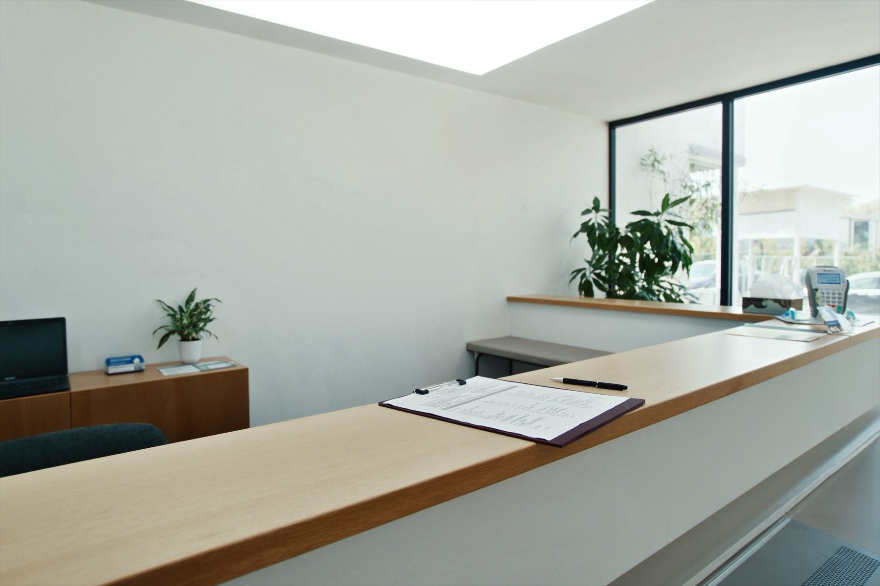 The front counter of the suite: a long light wood reception desk with a clipboard and pen set out on it, a plant, a desk phone and a window looking onto the parking lot.