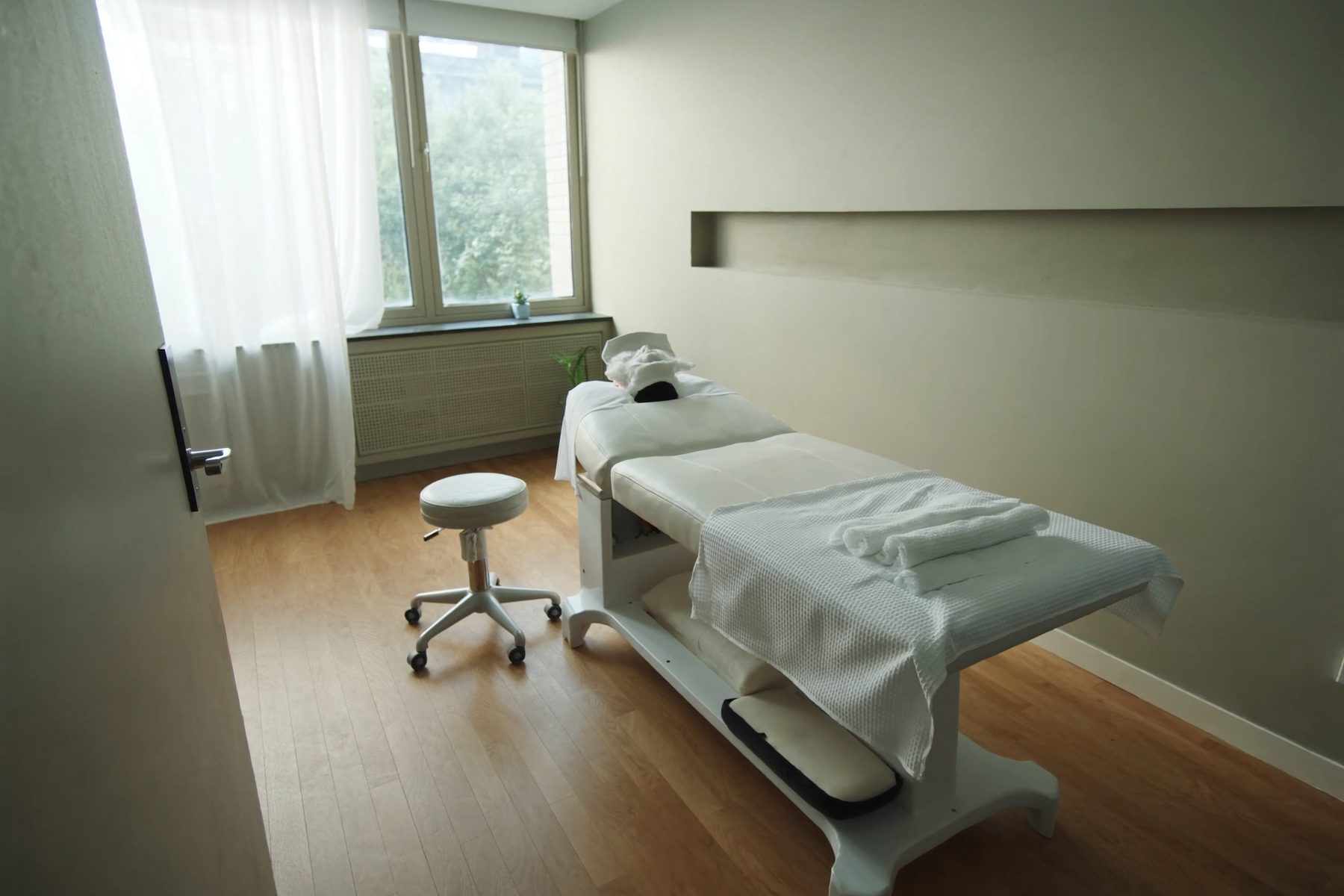 A treatment room dressed for the next appointment: one padded table under fresh white linens with a face cradle, a bolster and a rolling stool, lit by a curtained window.