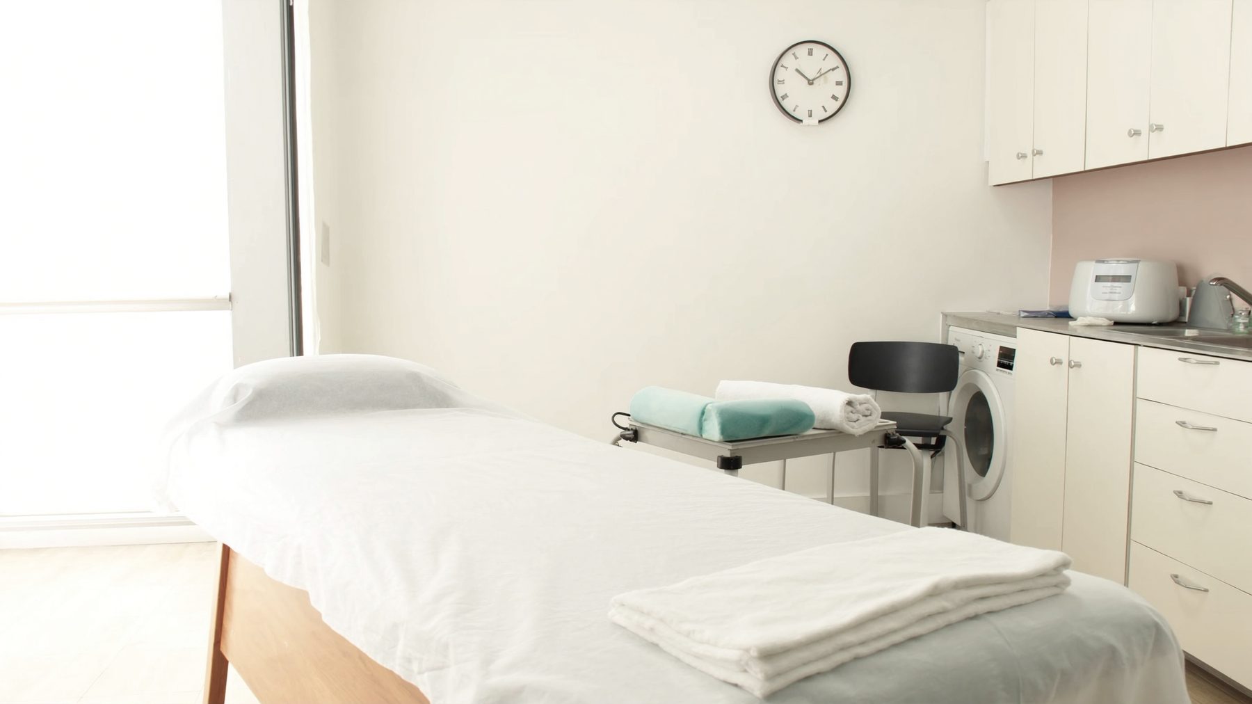 A treatment table made up along its length with a taut white sheet, a folded blanket at the foot, and bolsters and rolled towels waiting on a cart beside it under a wall clock.